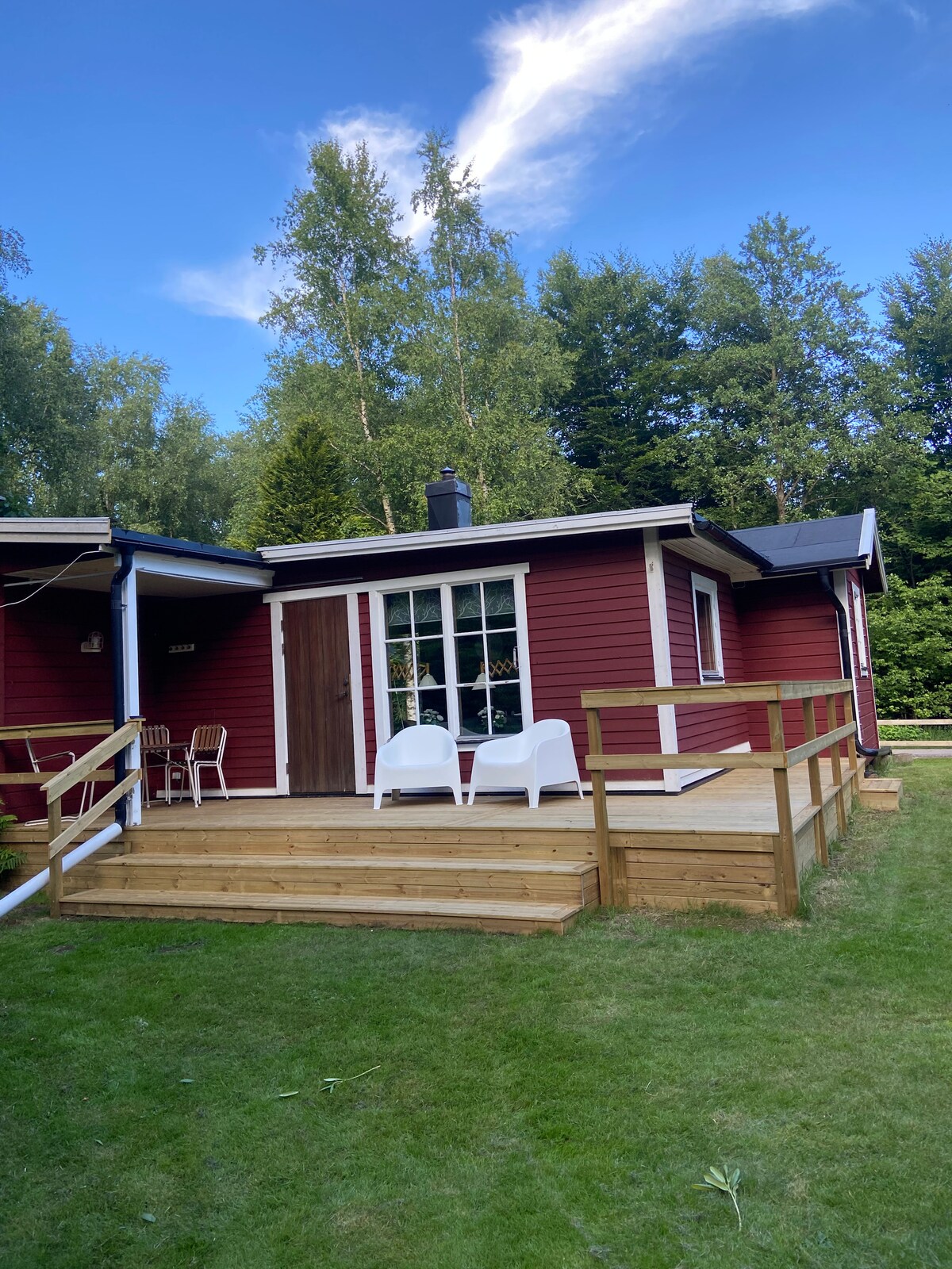 A red cottage with white trim is shown, featuring a wooden deck and a small set of white chairs. A large window reflects natural light from the surrounding greenery. The peaceful, grassy area is framed by trees, enhancing the sense of privacy.