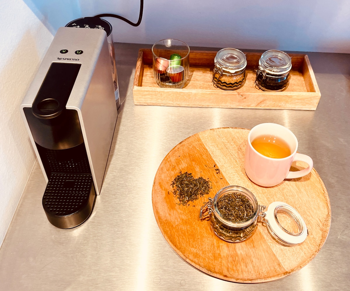A modern kitchen countertop displays a Nespresso coffee machine beside a wooden tray containing various tea jars and a cup filled with brewed tea. Loose leaf tea is scattered on the wooden cutting board, offering a cozy beverage setup.