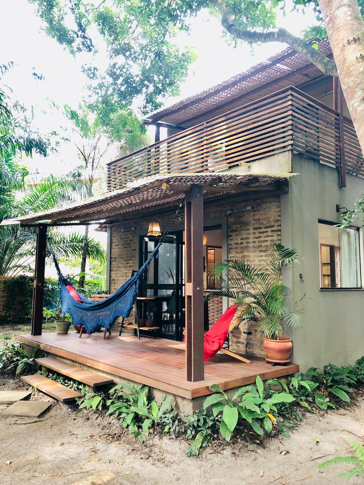 A charming exterior is showcased, featuring a wooden deck with a blue hammock and two red chairs. The building blends into the lush greenery, with large windows allowing for natural light. Decorative plants surround the entrance, enhancing the inviting outdoor space.
