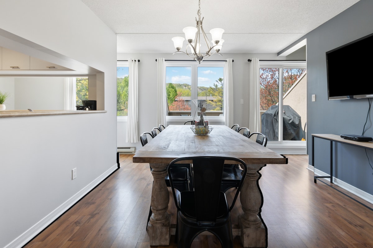 A spacious dining area is highlighted by a large wooden table with black chairs, seating eight comfortably. Natural light floods the space through large windows, offering views of the outdoors. A chandelier provides illumination overhead, while a flatscreen TV is mounted on a nearby wall.