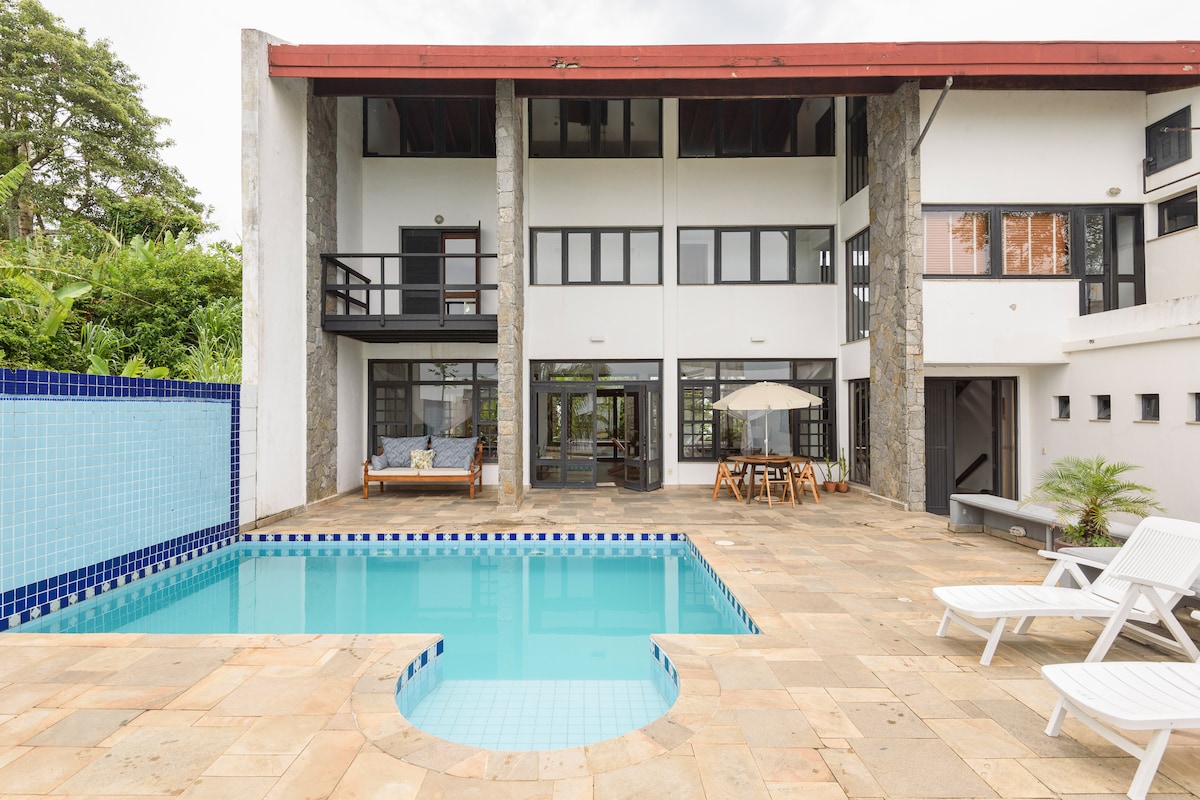 A spacious outdoor area features a private pool with a curved edge, surrounded by stone tiles. A dining table and chairs are positioned beneath an umbrella, while lounge chairs are available for relaxation. The two-story structure showcases large windows and an open layout, enhancing natural light.