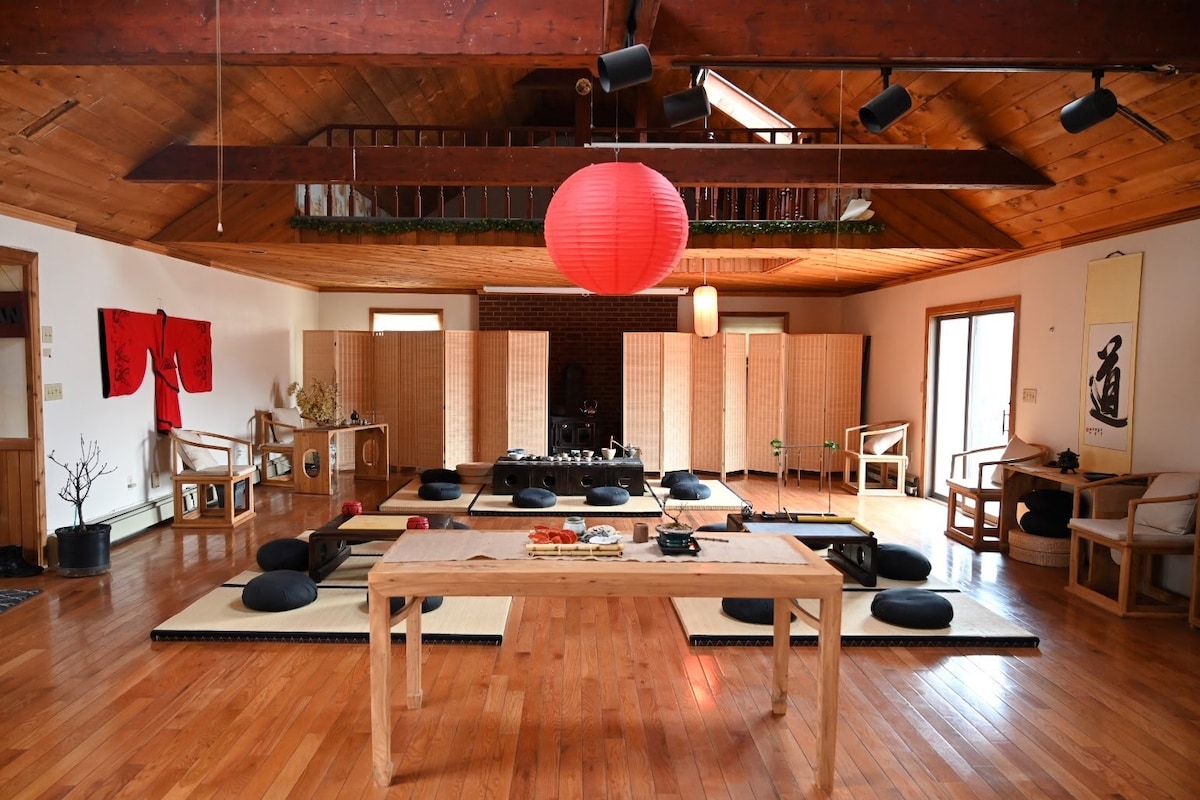 An open living space features wooden flooring with tatami mats and cushions arranged in a serene layout. A large, round paper lantern hangs from the ceiling, offering soft illumination. Wooden furniture and decorative screens delineate the area, creating a peaceful retreat for meditation or gatherings.