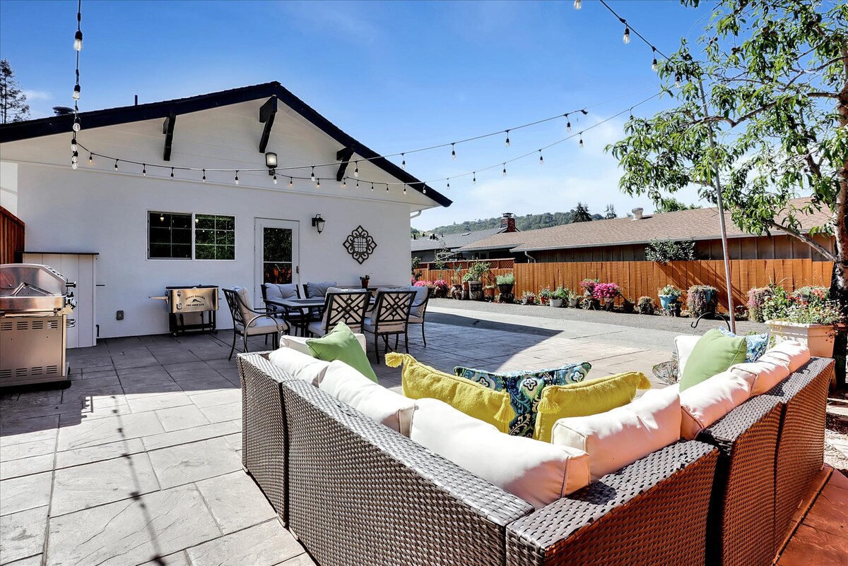 An outdoor patio area is presented with a modern sofa, adorned with vibrant cushions. Bistro lights hang overhead, illuminating the space. A barbecue grill is observed nearby, along with a dining table and chairs, surrounded by lush landscaping and a clear blue sky.