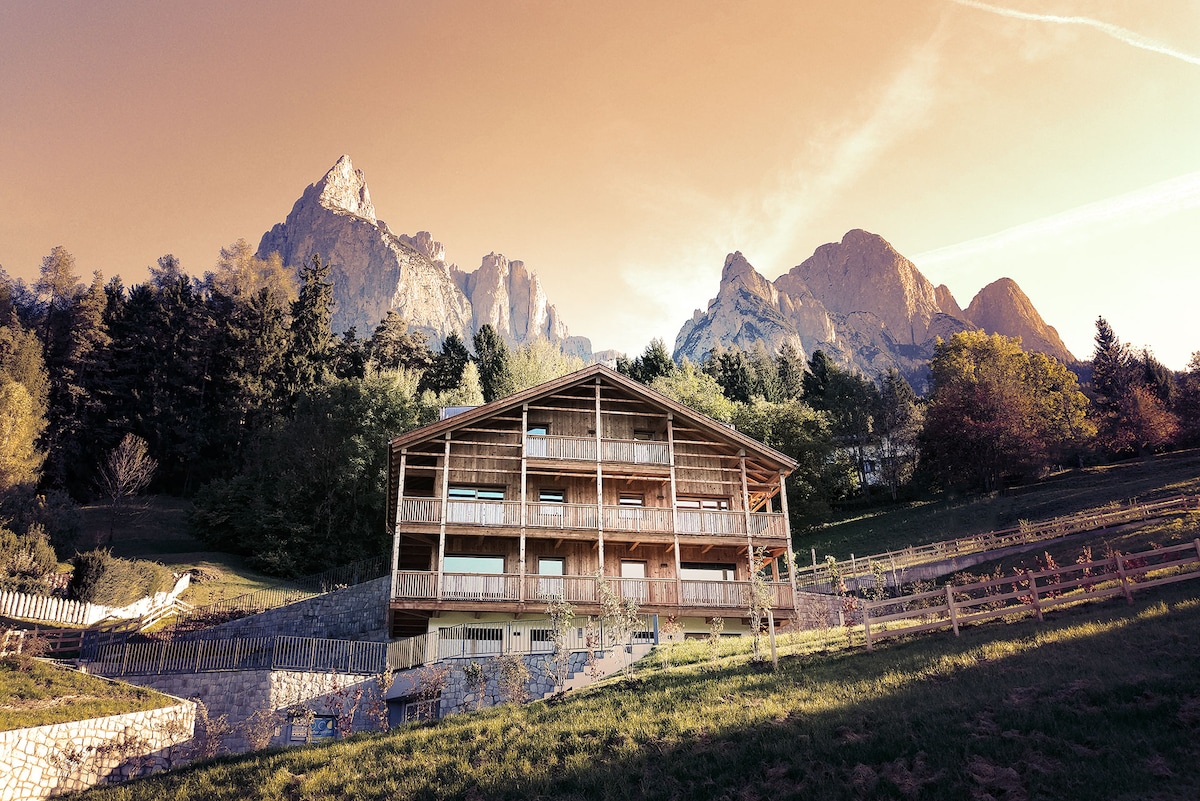 The chalet-style building is set against a backdrop of majestic mountains, with multiple balconies and large windows reflecting the natural surroundings. Lush greenery and terraced gardens enhance the landscape, providing a serene atmosphere beneath a warm-hued sky.