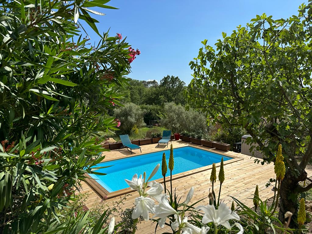 A refreshing swimming pool is framed by a wooden deck, surrounded by lush greenery and colorful flowers. Two lounge chairs are positioned beside the pool. Sunlight filters through the leaves, highlighting the serene garden setting.