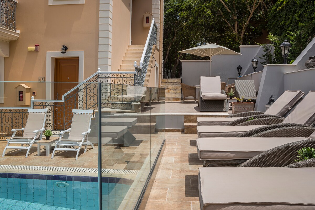 A sunlit outdoor area features a clear swimming pool bordered by light-colored lounge chairs with soft cushions. A large umbrella provides shade, while elegant lampposts line the pathway leading to nearby stairs.
