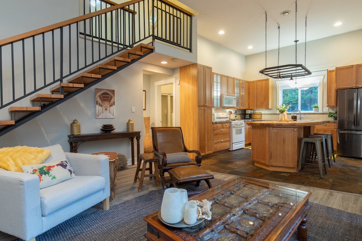 The open concept living area showcases a modern kitchen with wooden cabinetry and a centered island. Natural light floods the space through large windows. A cozy seating area features a light-colored armchair, a wooden coffee table, and decorative accents, with stair access to the upper loft area.