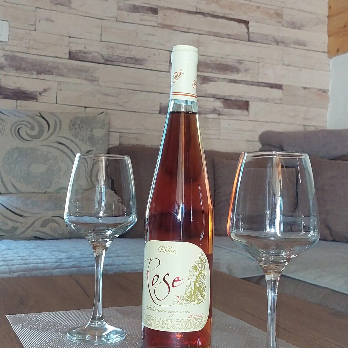 A bottle of rosé wine is positioned between two empty wine glasses on a textured table. The elegant label of the wine is clearly visible, and a comfortable sofa is seen in the background, contributing to a cozy setting.
