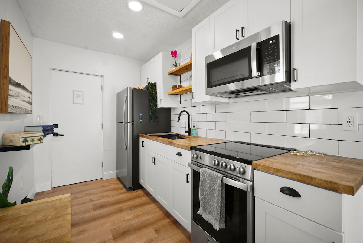 A modern kitchen features white cabinetry and open shelving. A stainless steel refrigerator stands alongside a gas range and an overhead microwave. Light wood countertops complement the sleek design, while potted plants add a touch of greenery. Natural light brightens the space.