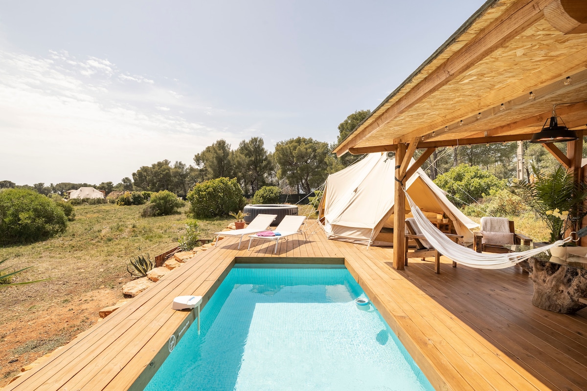 A private pool glistens beside a wooden deck, surrounded by greenery. A shaded area features a hammock and lounge chairs, while a spacious tent stands nearby. The open landscape of the countryside complements the serene outdoor setting.