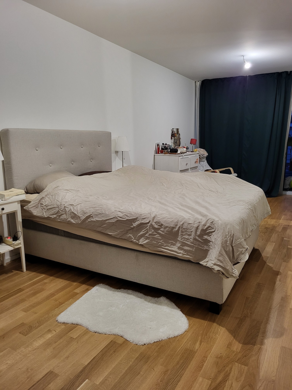 A spacious bed with a light-colored covering is centered in the room, complemented by a soft rug beneath. A bedside lamp and a small table are visible next to the bed. Dark curtains frame a window, while a white dresser holds personal items.