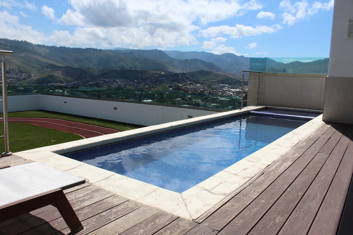 A sleek swimming pool is situated on a rooftop terrace, offering expansive views of the surrounding mountains and cityscape. The pool is bordered by a wooden deck and features clear blue water under a bright sky, creating a relaxing atmosphere for guests.
