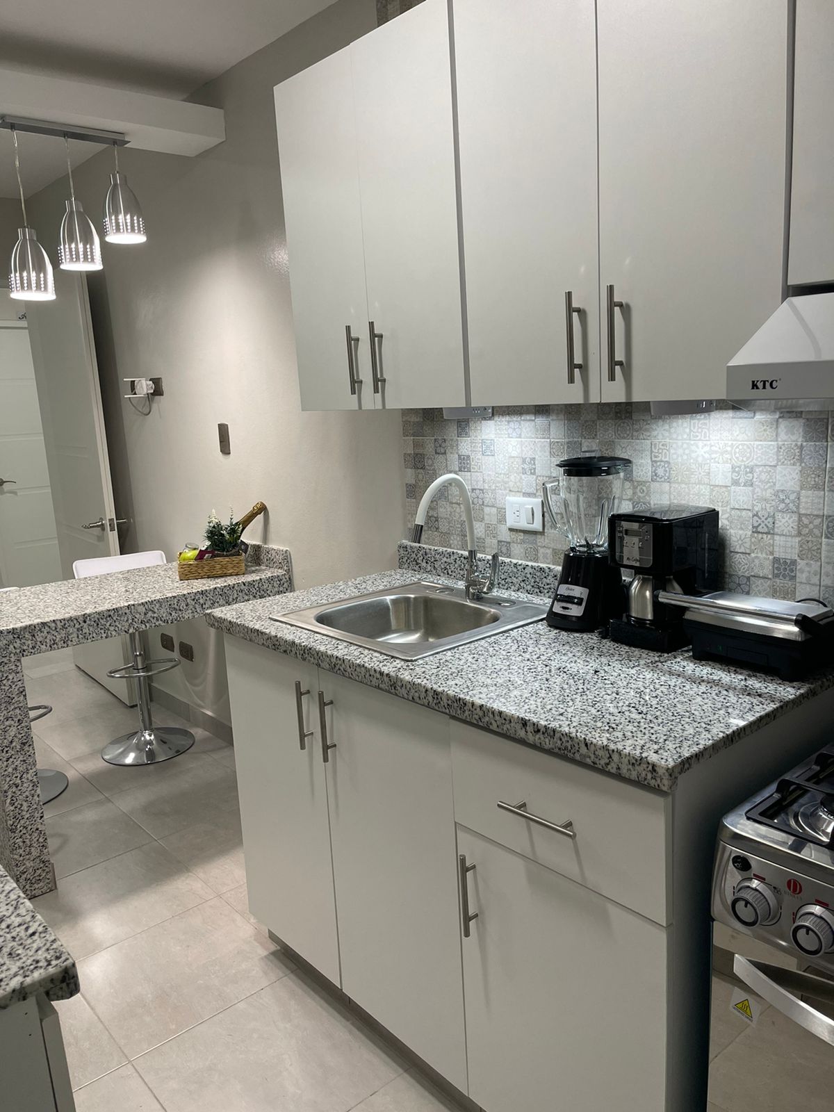 A modern kitchen is equipped with granite countertops and sleek white cabinetry. An under-cabinet light illuminates the workspace. A coffee maker and blender are placed on the counter. Bar stools are positioned at a small dining area, while pendant lights hang above for additional lighting.