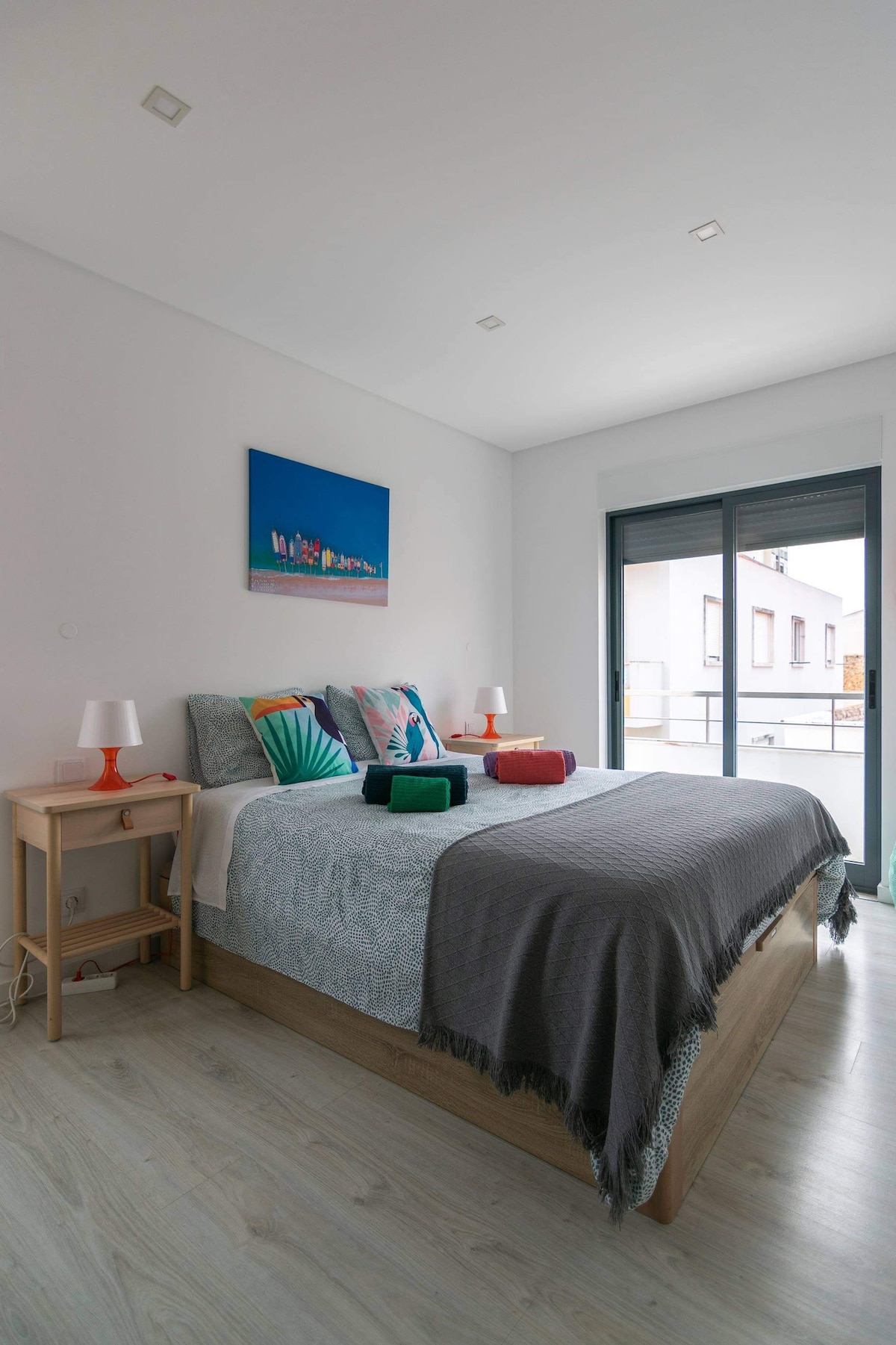 A well-lit bedroom features a spacious bed adorned with various decorative pillows and a textured throw. Nightstands with lamps flank the bed, providing additional light. A balcony door allows natural light to stream in, enhancing the calming atmosphere of the room.