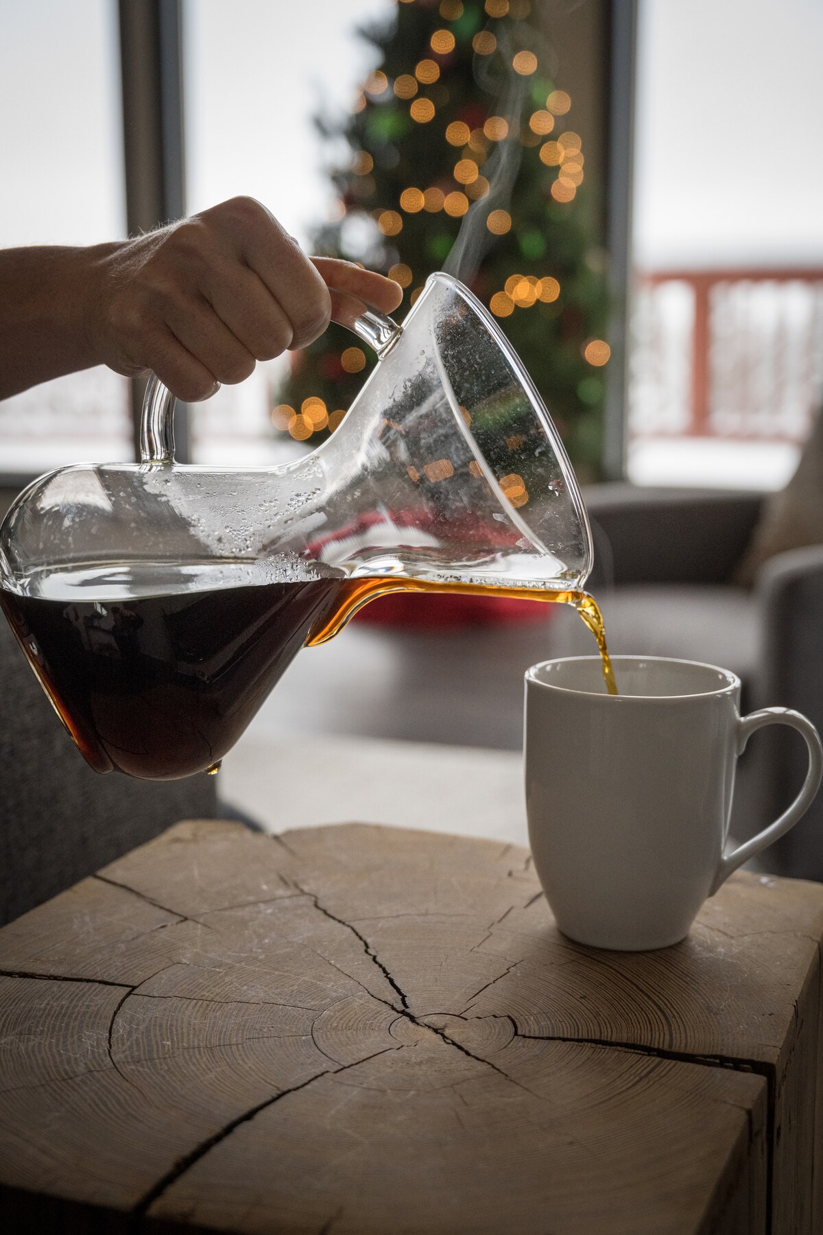 A hand pours coffee from a clear carafe into a white mug, with a wooden table in the foreground. Out of focus in the background, a Christmas tree decorated with warm lights adds a cozy atmosphere to the setting.