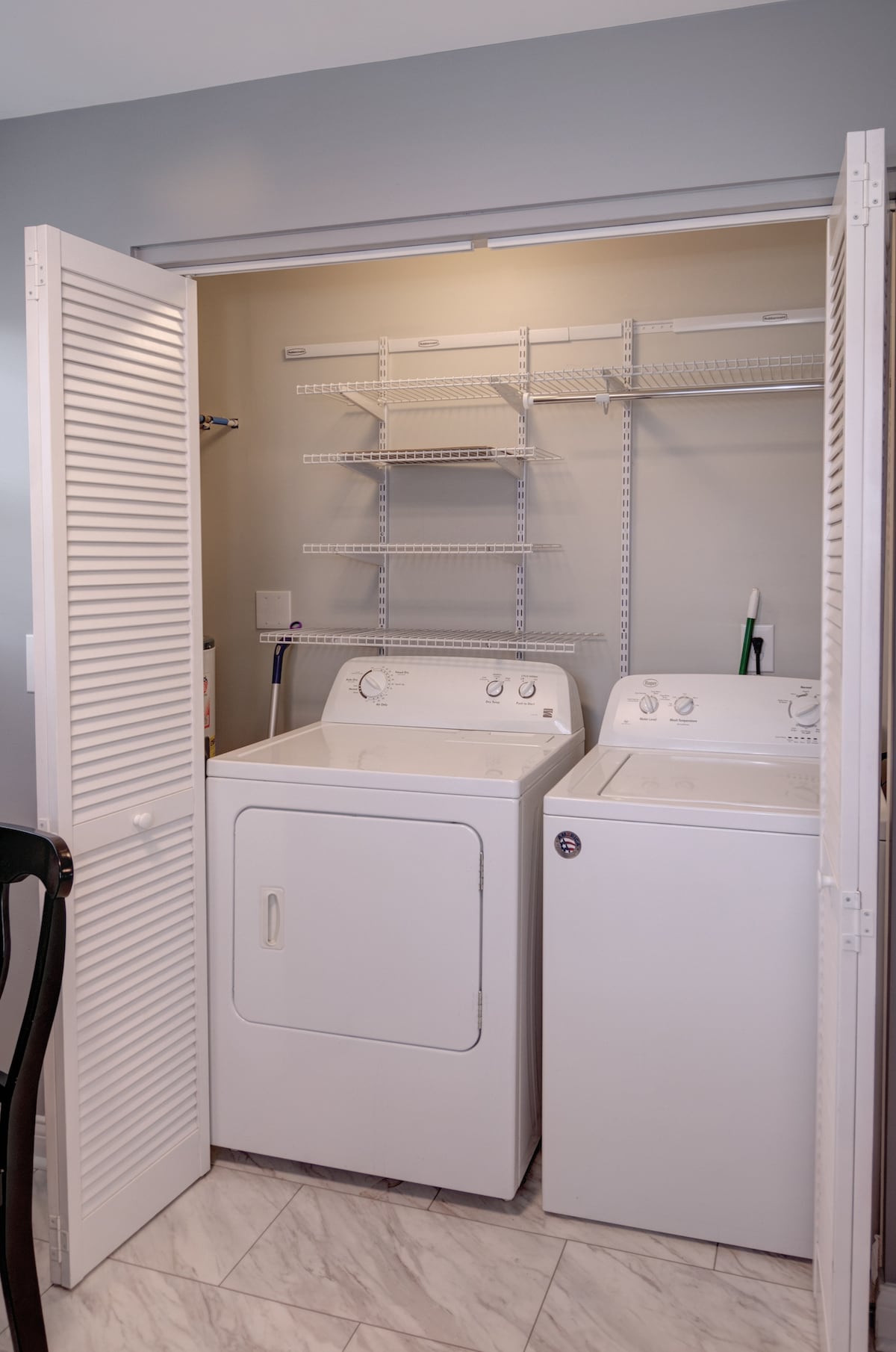 A compact laundry area features a stacked washer and dryer behind louvered doors. Above, wire shelving provides space for storage. The floor is finished with light-colored tiles, enhancing the practicality of this space.