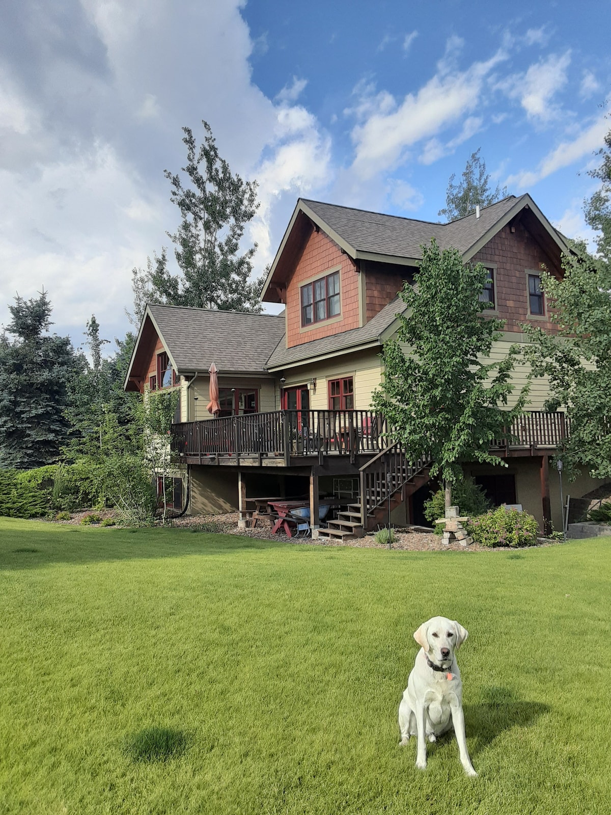 The exterior of a two-story home is framed by well-maintained landscaping and a spacious green lawn. A friendly dog sits on the grass, while a deck adorned with seating and shade is visible on the upper level, surrounded by trees.