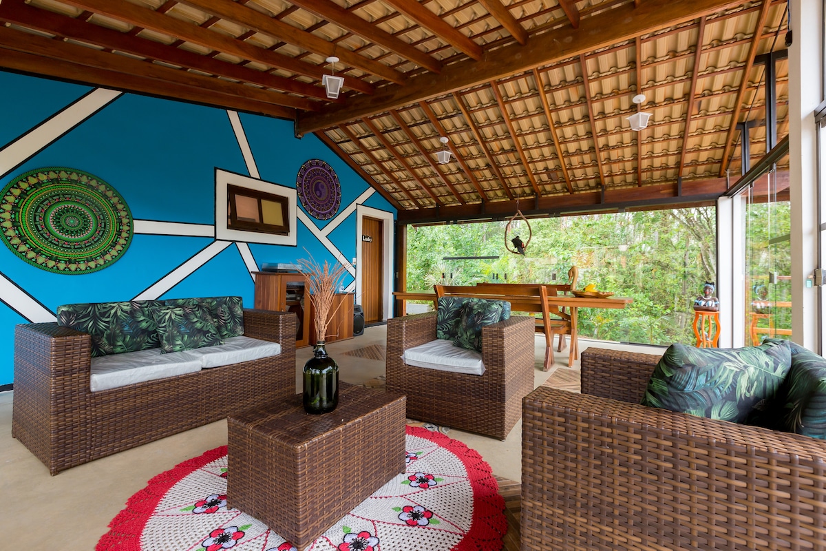 An inviting living area features a woven seating arrangement with comfortable cushions, complemented by a decorative circular rug. Vibrant blue walls adorned with patterned art enhance the space, while large windows provide views of the surrounding greenery.