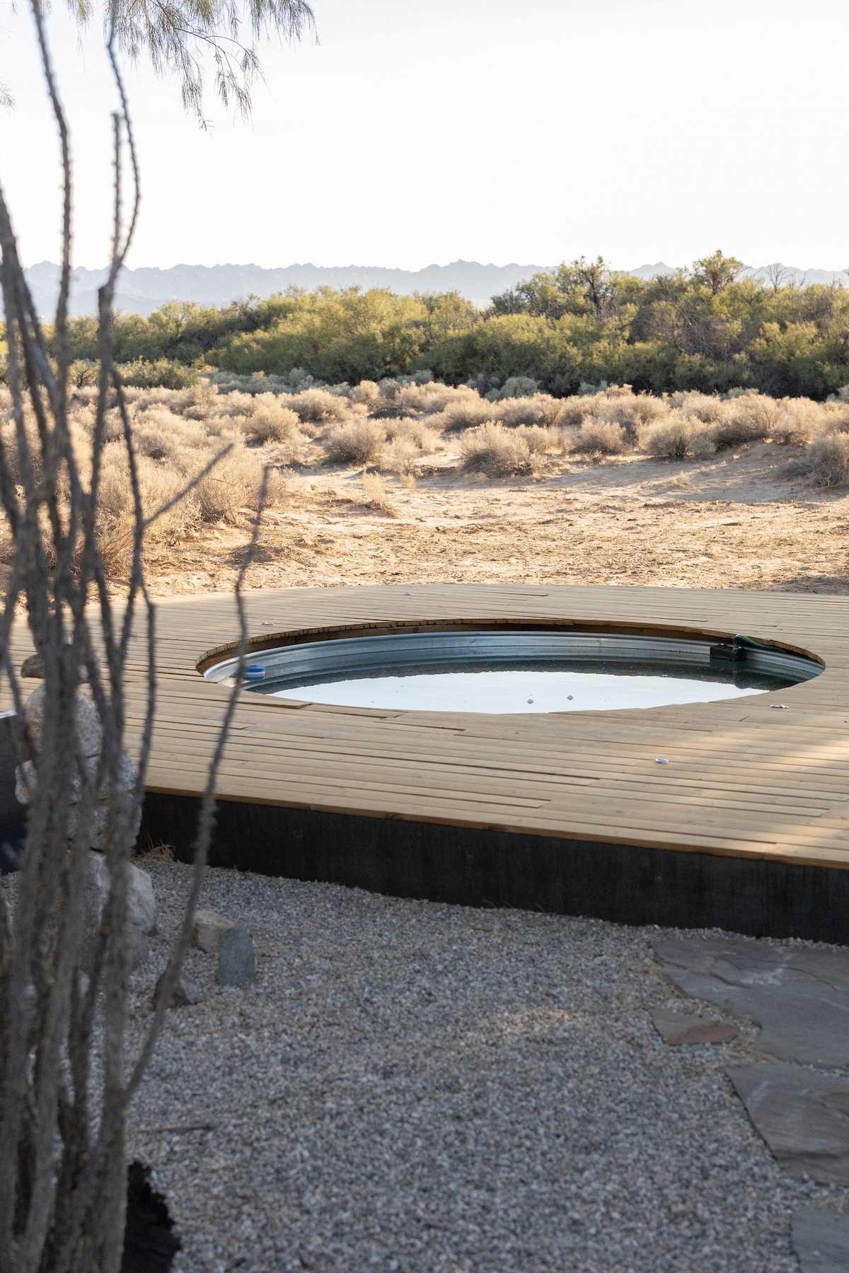 A circular wood deck surrounds a hot tub, which is nestled into the natural landscape. The area is framed by desert flora, and the scene features expansive views of the distant hills and shrubs, providing a tranquil outdoor experience.
