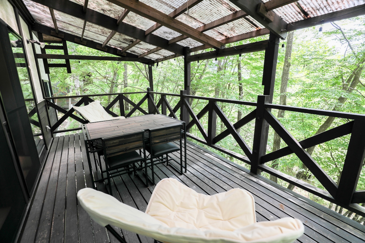 An elevated wooden deck is surrounded by lush greenery, providing a serene outdoor space. A rectangular dining table with matching chairs sits centrally, complemented by two lounge chairs. The slatted roof allows natural light to filter through while offering a shaded environment.