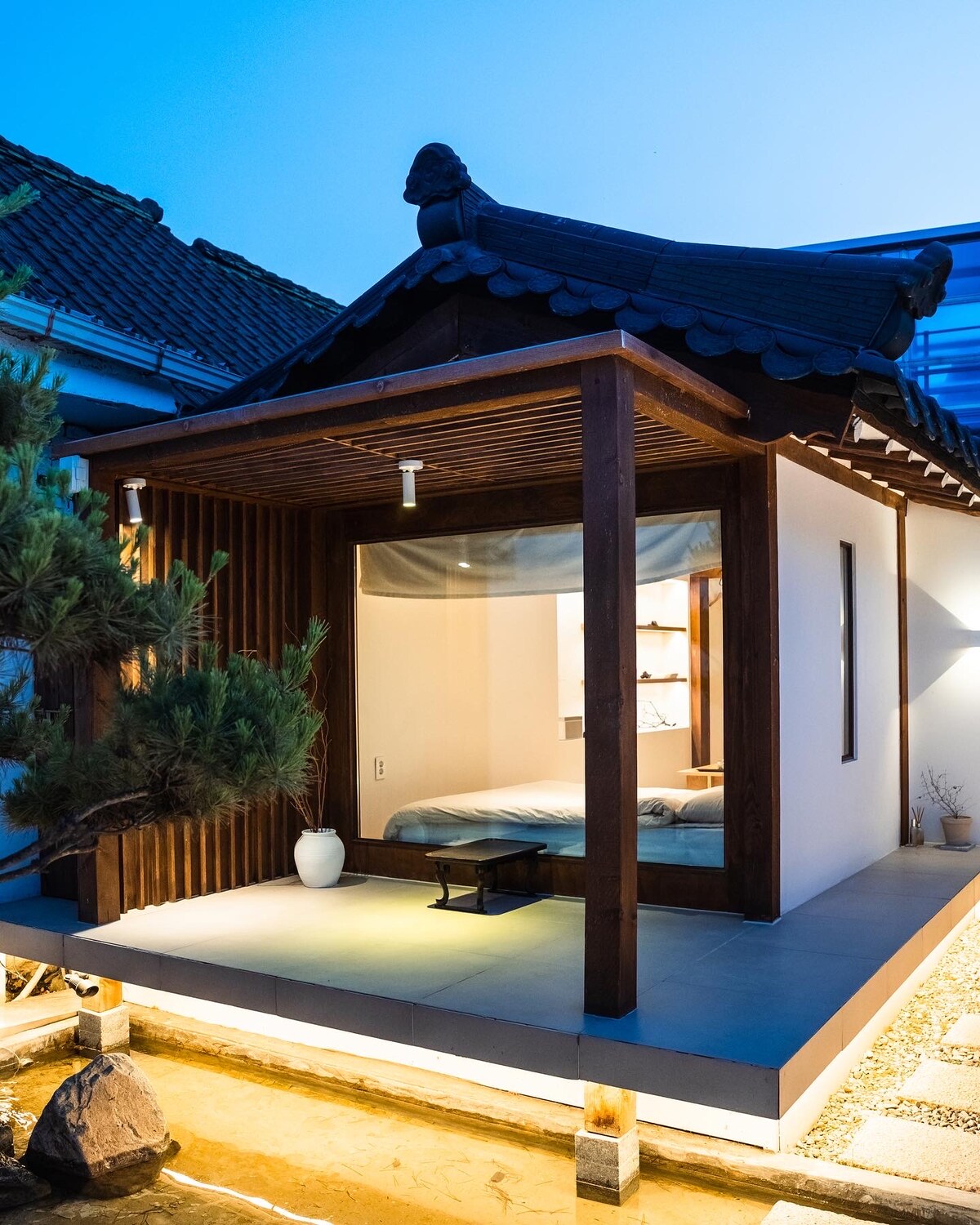 The exterior of the traditional Hanok-style house is illuminated by soft lighting, highlighting its wooden structure and unique roof design. A small patio area features a minimalist table, while a tranquil pathway leads to the entrance. A serene ambiance is created by surrounding nature.