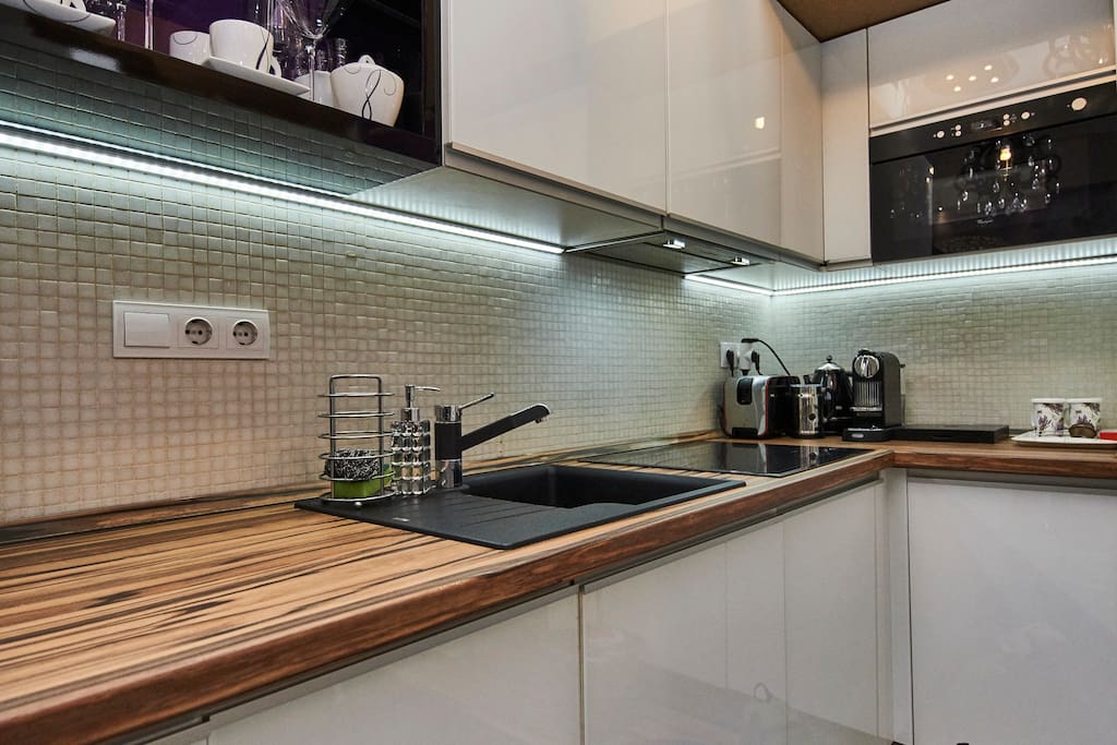 A modern kitchen space features a sleek wooden countertop with a black sink and chrome faucet. Above, cabinetry is illuminated by LED lights, highlighting dishes and glassware. Kitchen appliances, including a coffee maker and kettle, are neatly arranged on the countertop.