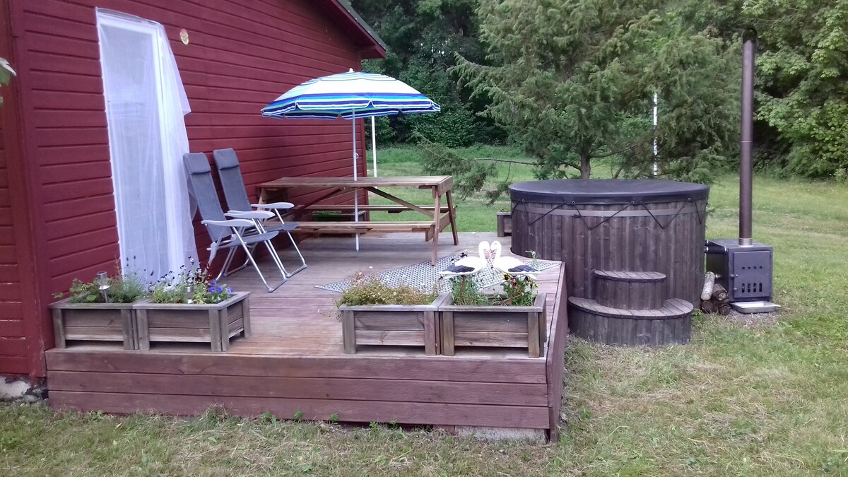 A wooden deck features a picnic table under a striped umbrella and two lounge chairs. Planters with flowers add greenery, while a wooden hot tub and a small wood stove enhance the outdoor setting surrounded by trees.