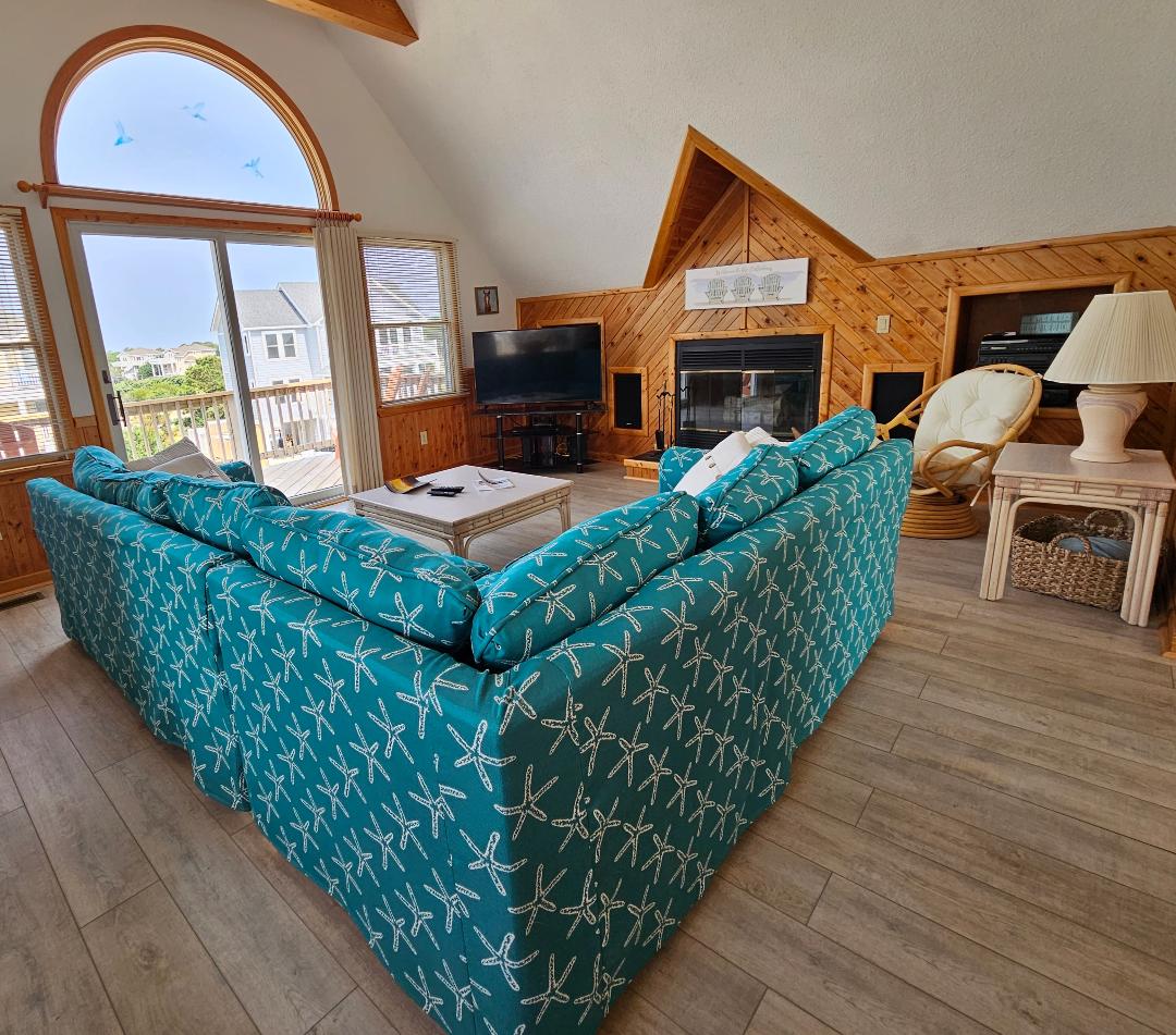 A spacious living area features a large sectional sofa upholstered in a starfish pattern, offering ample seating. A flat-screen TV is positioned on the opposite wall, with a fireplace nearby. Natural light fills the room through large windows that overlook the outdoors.