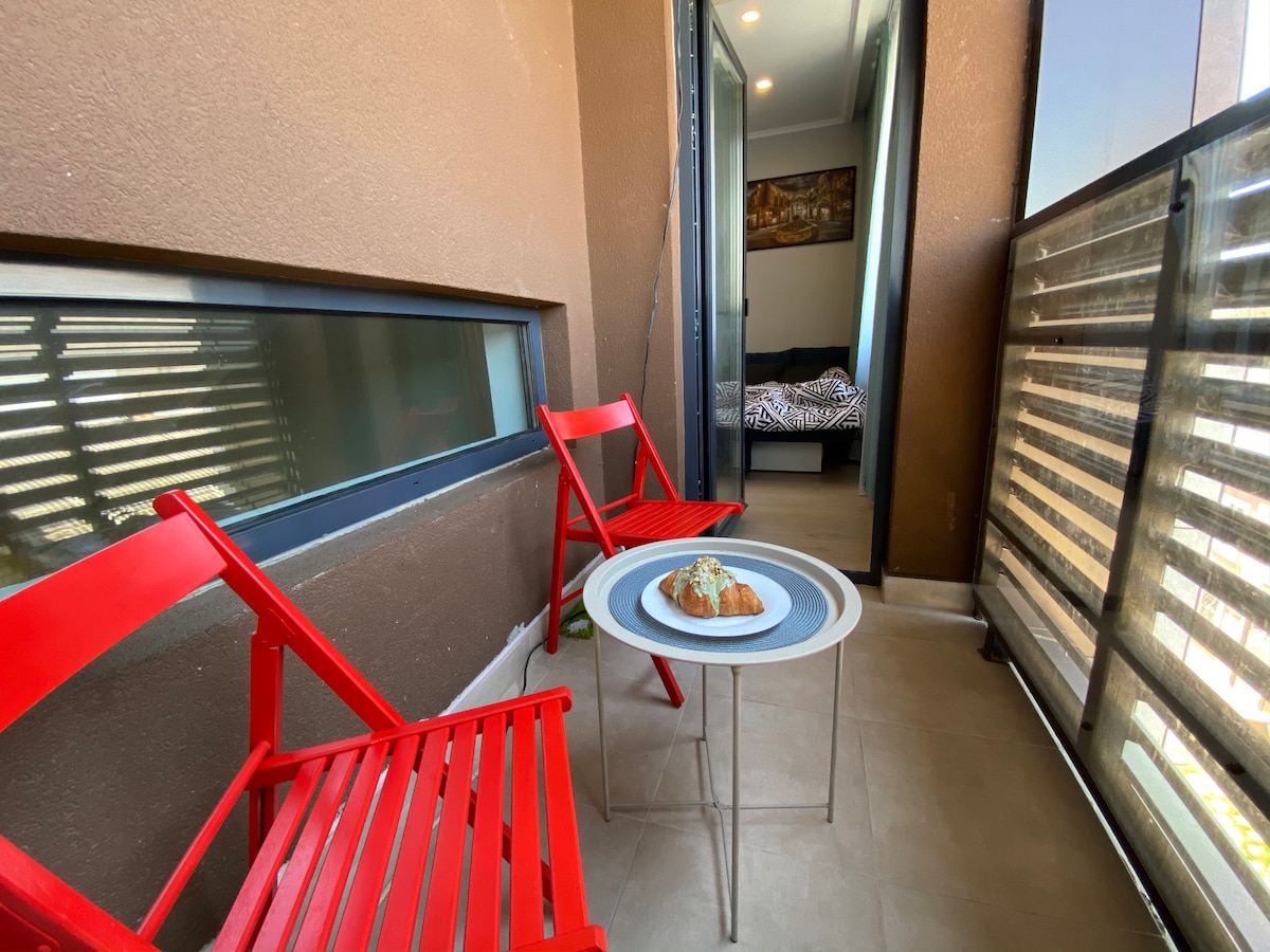 A balcony area is shown featuring two red chairs and a small round table, which holds a pastry. Sunlight is reflected from nearby large windows, enhancing the space's brightness. The background reveals a glimpse of a bedroom with dark bedding.