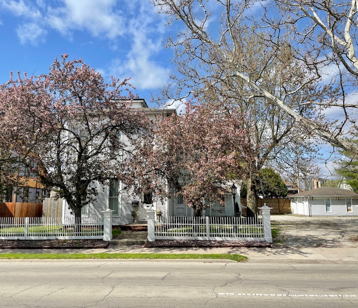 The exterior of the Phillips-Johnson House is showcased, framed by blooming trees and a white picket fence. The two-story building features a mixture of windows and a front porch, with a view of the spacious parking area behind.