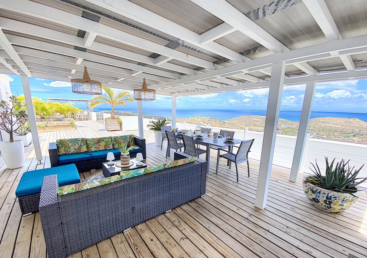 An outdoor terrace is furnished with a comfortable seating area featuring a sectional sofa with colorful cushions. A dining table with chairs accommodates several guests, and a scenic view of the turquoise waters is visible from the space. Light fixtures hang from the ceiling, enhancing the ambiance.