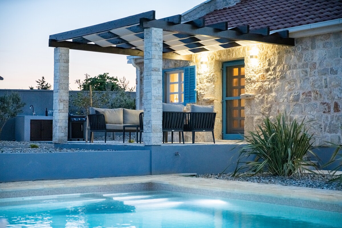 A stone patio features a comfortable seating area beneath a shaded pergola, showcasing a modern design with dark wicker chairs and neutral cushions. The tranquil private pool reflects soft evening lights, creating a calm environment surrounded by low-maintenance landscaping and a serene backdrop.