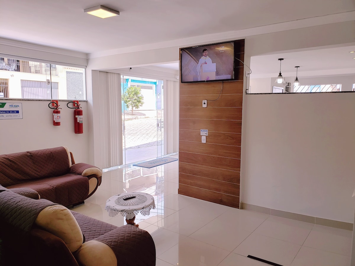 A welcoming lobby area is presented, featuring a comfortable seating arrangement with a distinctive couch beside a small table. A television is mounted on the wall, displaying a program. Large windows allow natural light to fill the space.