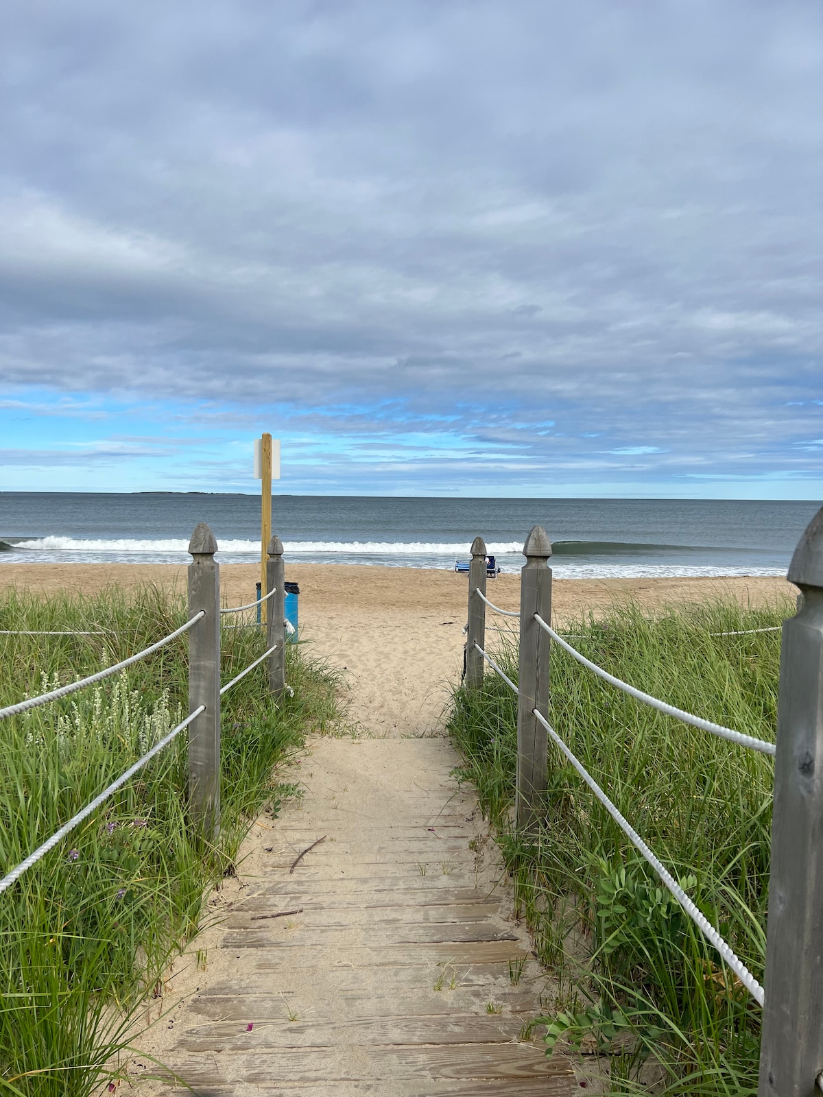 A sandy pathway leads towards the beach, bordered by tall grass on either side. Wooden posts with ropes guide the way to the shore, where gentle waves can be seen lapping at the coastline under a partly cloudy sky.