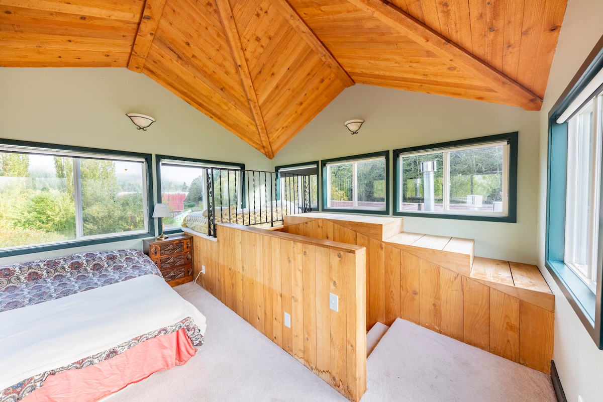 A spacious bedroom suite features a vaulted wooden ceiling and ample windows offering views of the surrounding landscape. A comfortable bed is complemented by a wooden chest of drawers. A built-in seating area provides additional space, highlighted by natural light filtering through the windows.
