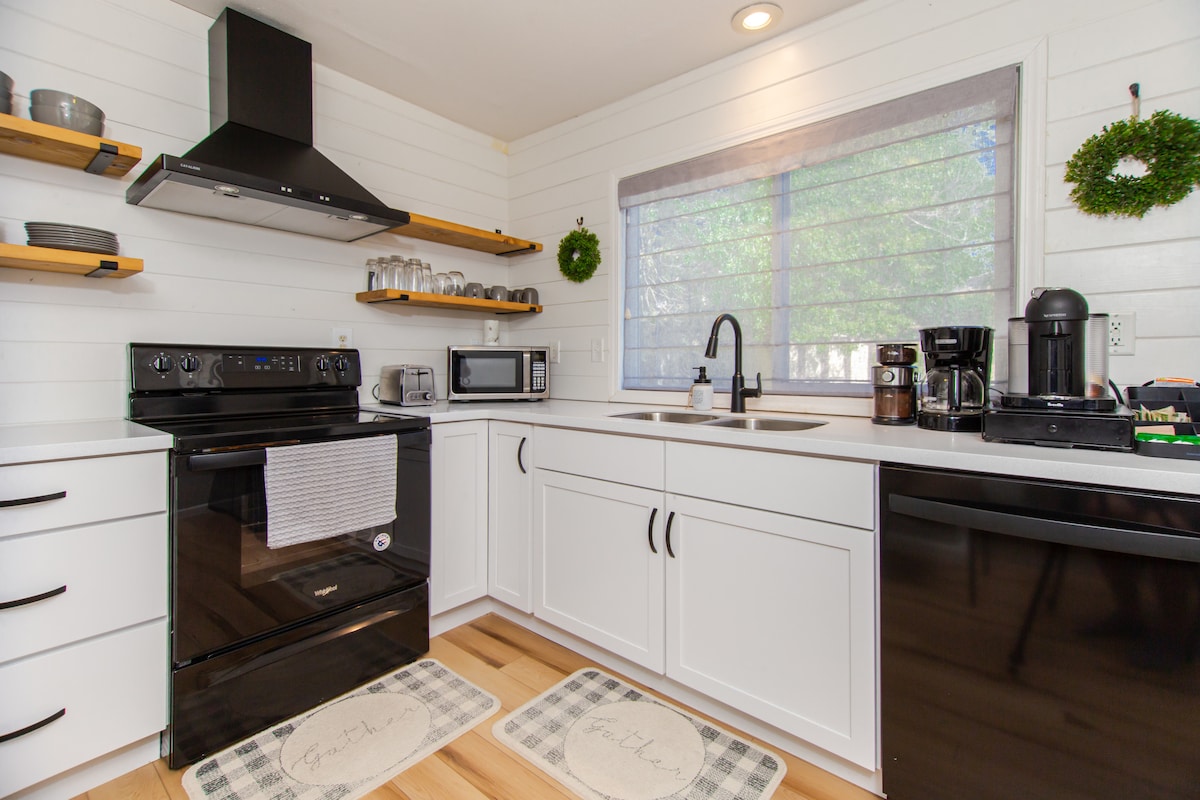 The modern kitchen is equipped with black appliances, including an oven and a microwave. White cabinets provide storage, with open shelves displaying dinnerware. A window allows natural light to brighten the space, enhanced by greenery decorations. The area features light wood flooring and patterned rugs.
