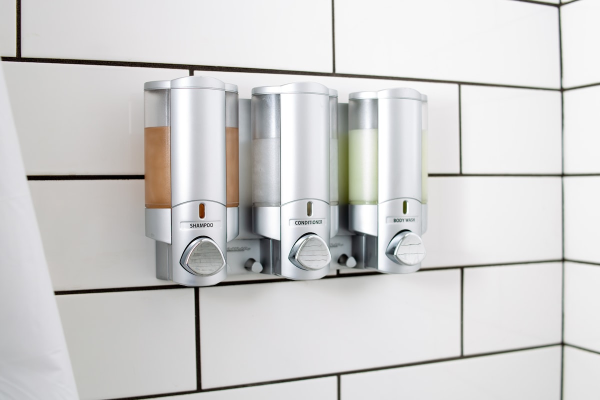 Three dispensers arranged on a wall feature shampoo, conditioner, and body wash. The sleek, silver containers are mounted on a tiled wall with a clean white and black grid pattern, providing easy access to essential bathing products.