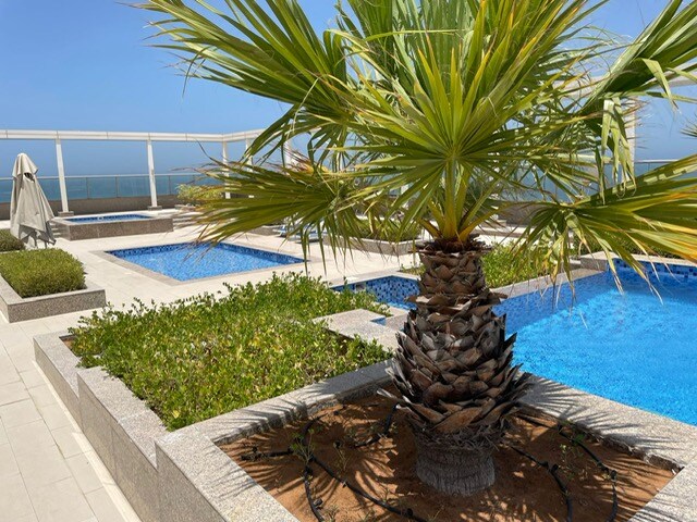 A rooftop area features three inviting swimming pools surrounded by landscaped greenery. A large palm tree stands prominently in the foreground, adding a tropical touch. Sun loungers are arranged nearby, providing a relaxing atmosphere with ocean views in the background.