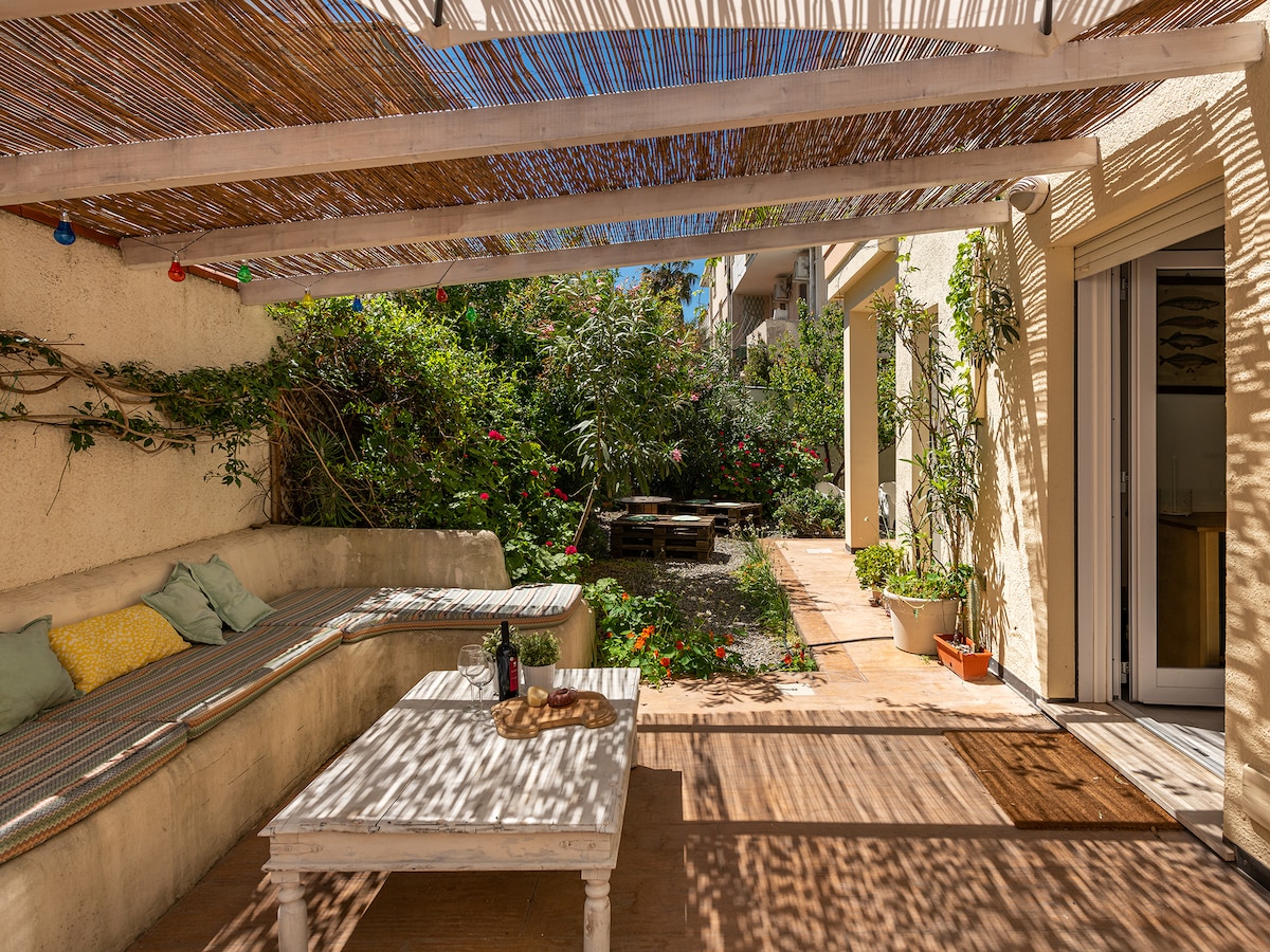 A shaded outdoor seating area features a built-in bench with soft cushions and a rustic wooden table. Lush greenery surrounds the space, complemented by sunlight filtering through a decorative overhead covering. A doorway leads into the interior of the home, enhancing the connection to nature.