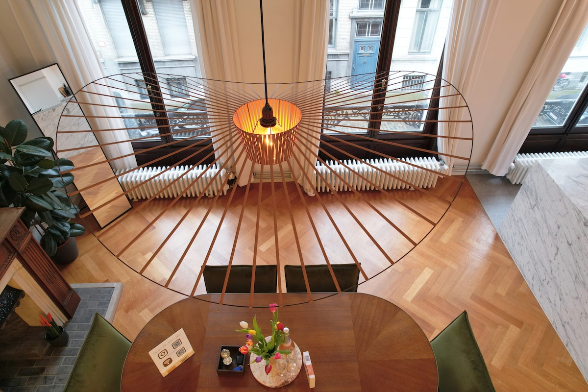 A modern dining area features a unique pendant light suspended above a round wooden table surrounded by green chairs. Large windows allow natural light to fill the space, highlighting the elegant herringbone floor pattern and minimalist decor.