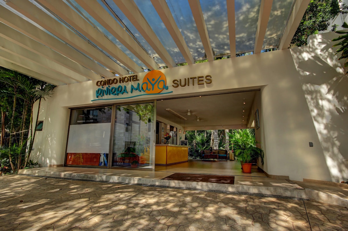 The entrance of the Condo-Hotel is framed by a modern overhang, providing shade. Lush tropical plants are visible around the pathways, contributing to a welcoming atmosphere. A reception area is seen through the expansive glass doors, emphasizing openness and accessibility.