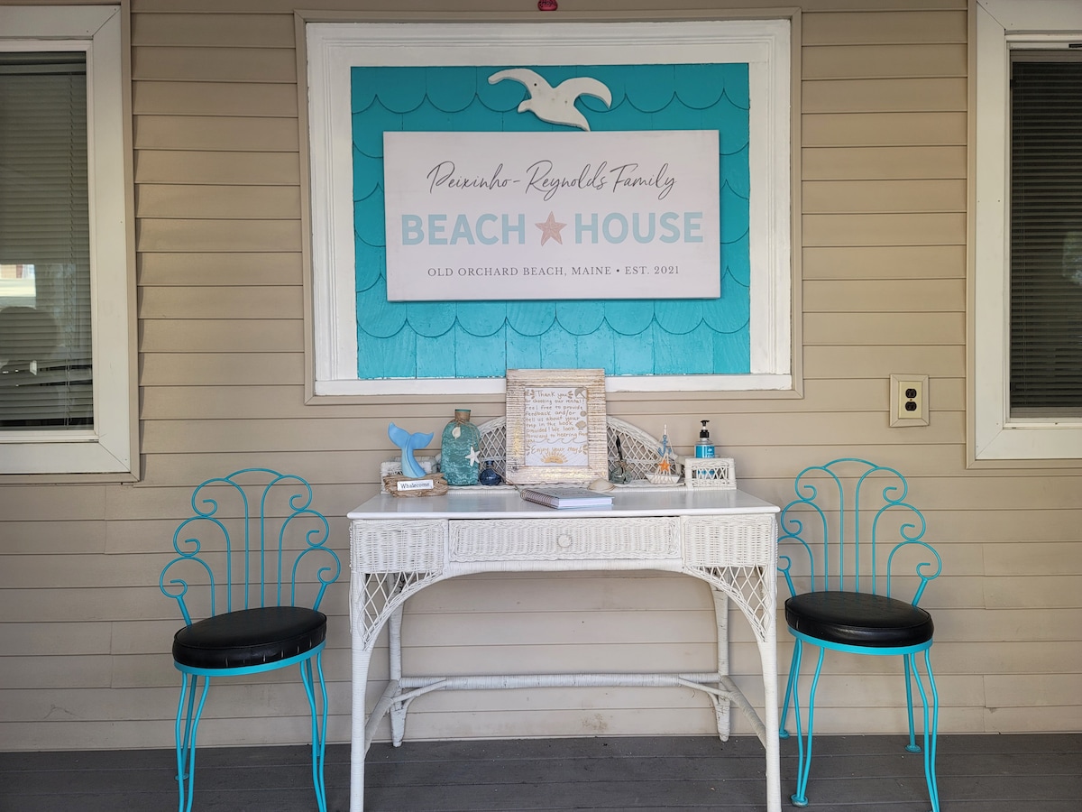 A welcoming entryway features a decorative sign that reads 'Beach House,' complemented by a coastal-themed design. Two blue chairs are positioned by a white table, which holds decorative items and a beach-themed ornament. Natural light filters through nearby windows.