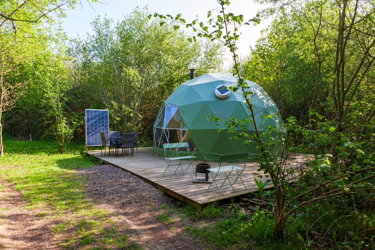 A geodesic dome is set on a wooden deck amidst greenery, showcasing a compact outdoor seating area with four chairs and a side table. A solar panel is positioned nearby, blending with the natural landscape. Sunlight filters through surrounding trees, enhancing the serene environment.