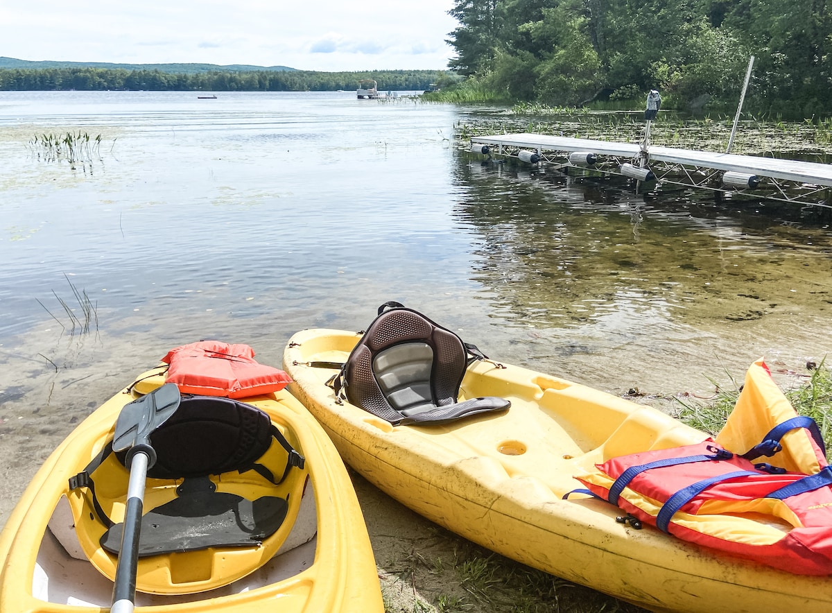 Two yellow kayaks rest on a sandy shoreline near a calm lake. A wooden dock extends into the water, with lush greenery surrounding the area. Life jackets are neatly arranged on the kayaks, hinting at opportunities for outdoor adventures.