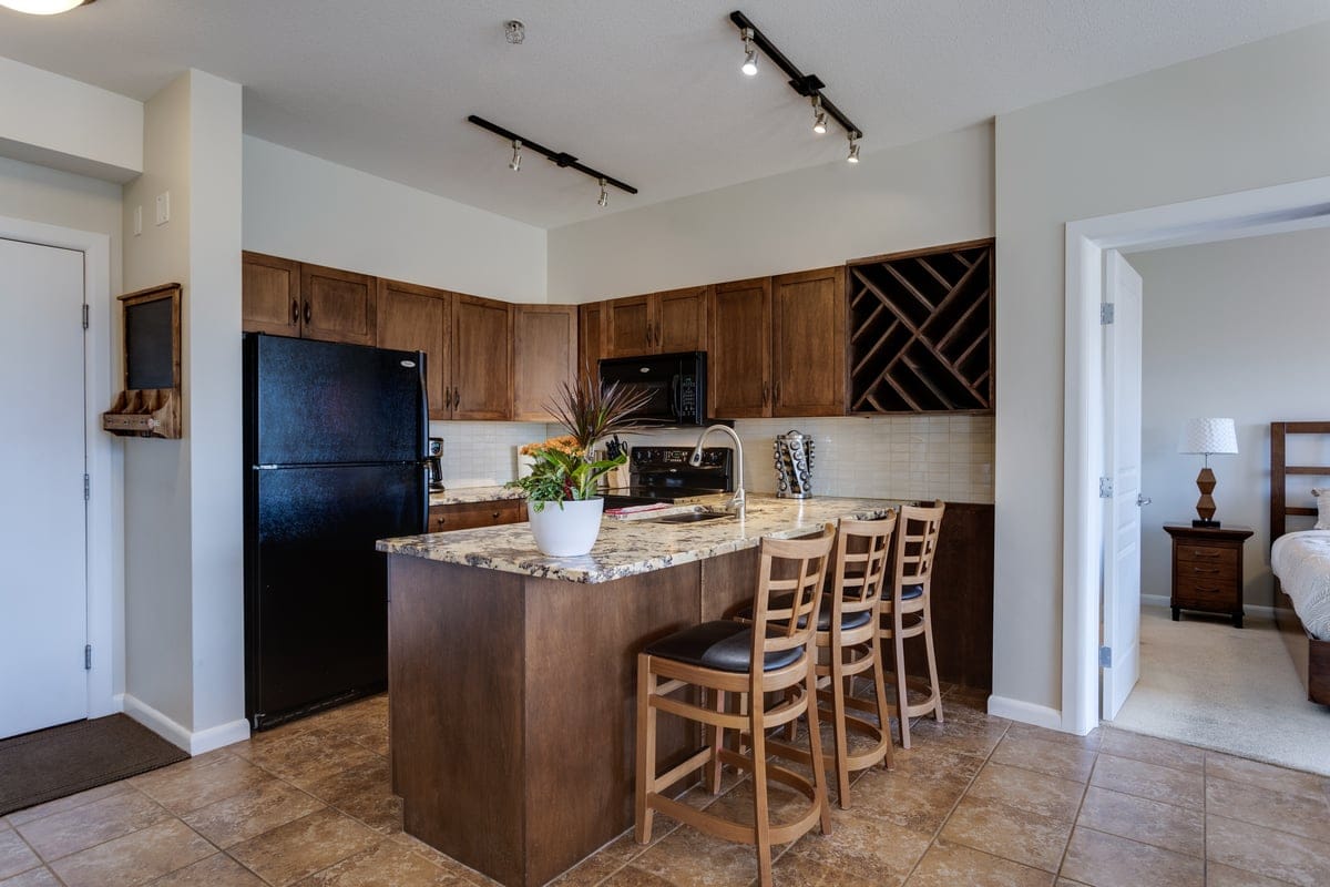 A modern kitchen is shown with dark wood cabinetry and granite countertops. A black refrigerator and microwave provide essential appliances. Four wooden barstools are arranged around a kitchen island, while natural light enters from a nearby entryway.