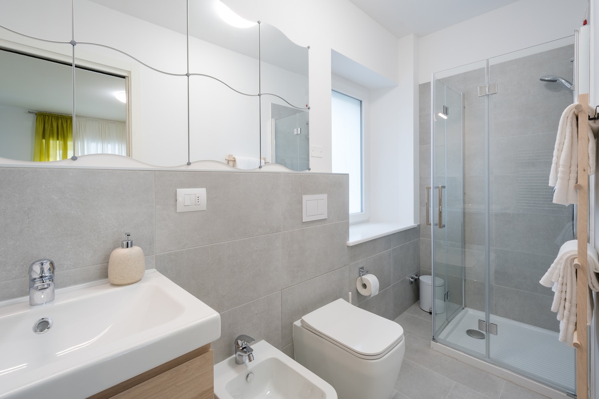 The modern bathroom features a glass shower stall and a sleek vanity with a sink. A spacious window allows natural light to brighten the space, complemented by a large mirror above the sink. Soft towels are neatly arranged for convenience.