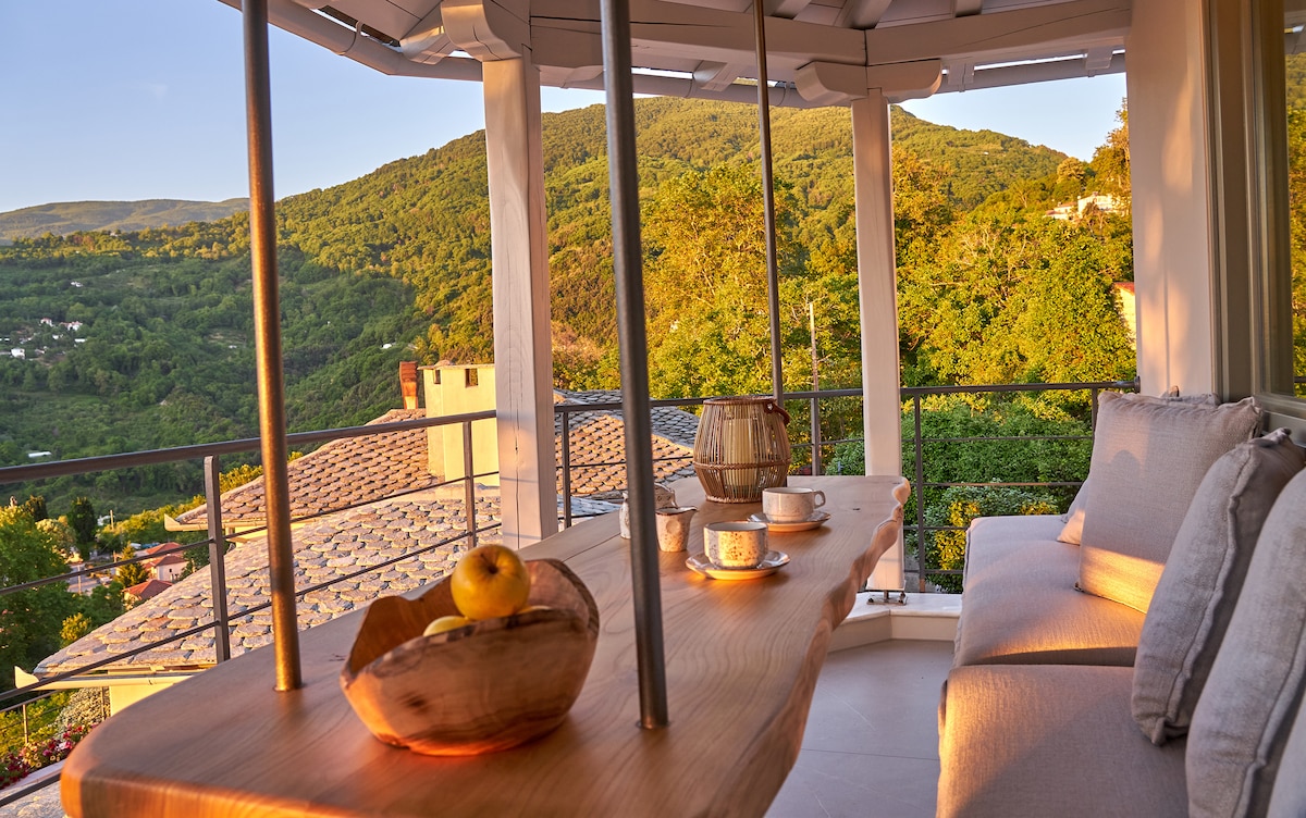 An outdoor balcony is featured, showcasing a wooden table with a bowl of fruit and two cups. Comfortable seating is arranged around the table, and the surrounding mountains are visible in the background, illuminated by warm sunlight.
