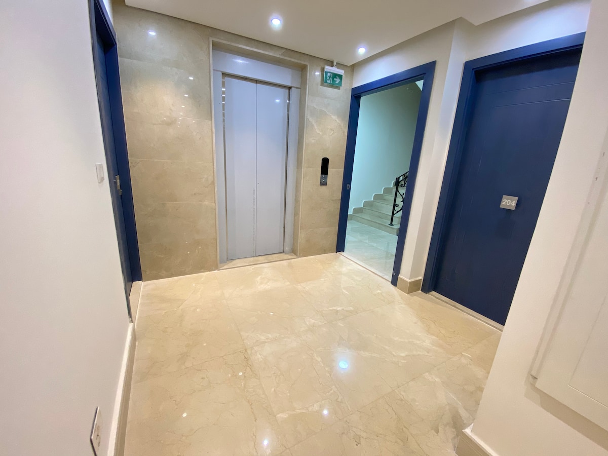 A clean and modern hallway features glossy marble flooring and light-colored walls. An elevator is positioned in the center, with two doors leading to private spaces on either side. Soft lighting enhances the spaciousness of the area, creating an inviting entrance.
