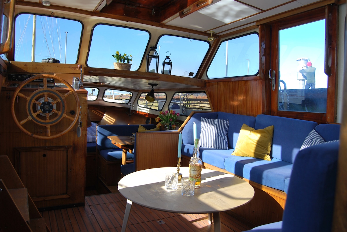 A spacious living area features wooden paneling and large windows that provide ample natural light. Comfortable blue seating surrounds a round table, with decorative items placed atop. A ship's wheel adds a nautical touch, enhancing the cozy atmosphere onboard.