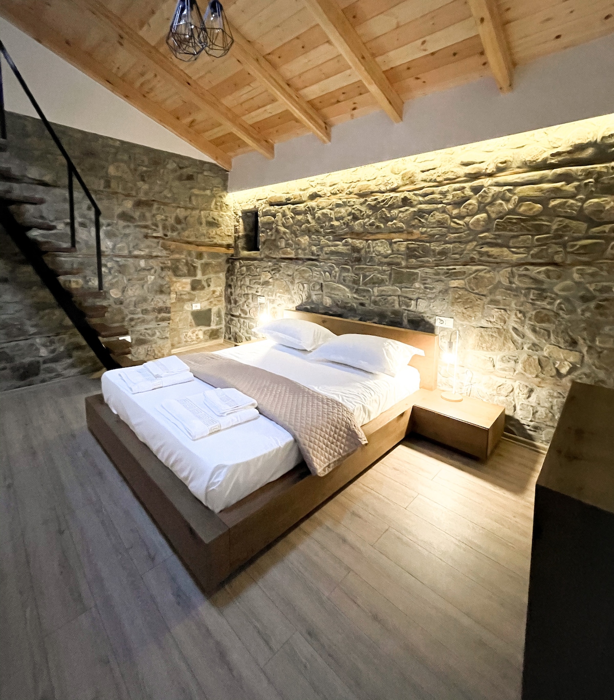 A spacious room is presented with a wooden ceiling and stone wall accents. A large bed is positioned at the center, accompanied by minimalistic bedside tables. Soft lighting creates a serene ambiance, and a staircase is visible in the background, leading to an upper level.
